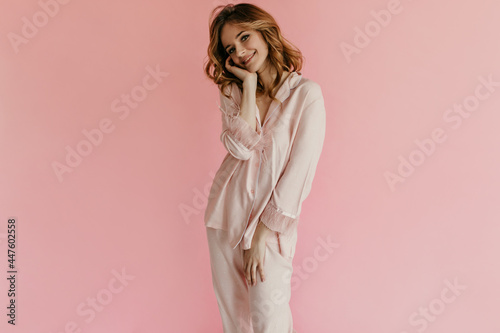 Cute young woman with wavy blond hair in stylish light pink suit looking into camera and smiling on isolated background..