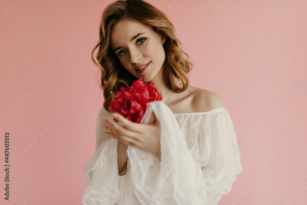 Cute curly girl with delicate makeup enjoys smell of bouquets of red tulips on pink background. Beautiful lady in white shirt looking into camera.. Cute curly girl with delicate makeup enjoys smell of bouquets of red tulips on pink background. Beautiful lady in white shirt looking into camera..