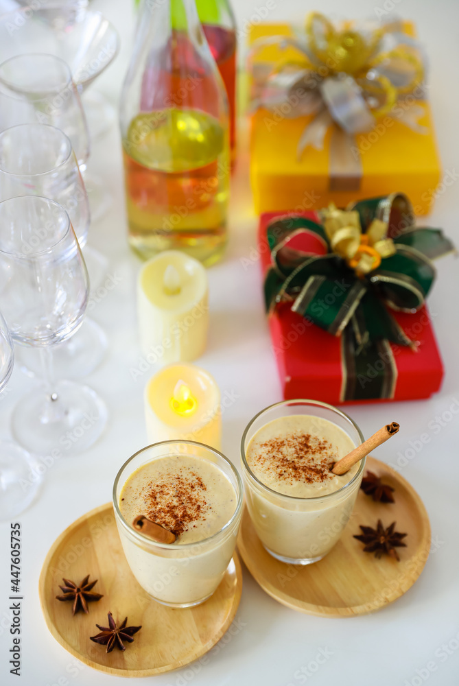 Top view shot of two sweet creamy alcoholic eggnog drinks with gingerbread on wooden coaster with anise on white clothing table full of empty tall wine glasses liquor bottles and present gift boxes
