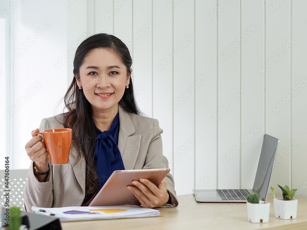 Asian female office manager with brown suit holding tablet computer in her hand. She smile happily at the taste of hot coffee. Morning work atmosphere in a modern office. Asian female office manager with brown suit holding tablet computer in her hand. She smile happily at the taste of hot coffee. Morning work atmosphere in a modern office.