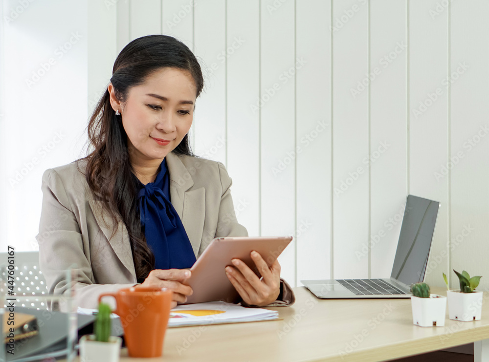 Asian female office manager with brown suit holding tablet computer in her hand. Morning work atmosphere in a modern office. Asian female office manager with brown suit holding tablet computer in her hand. Morning work atmosphere in a modern office.