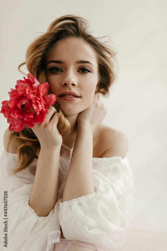 Portrait of young lady with curly blond hair in light modern blouse posing with pink beautiful flower on white background.. Portrait of young lady with curly blond hair in light modern blouse posing with pink beautiful flower on white background..