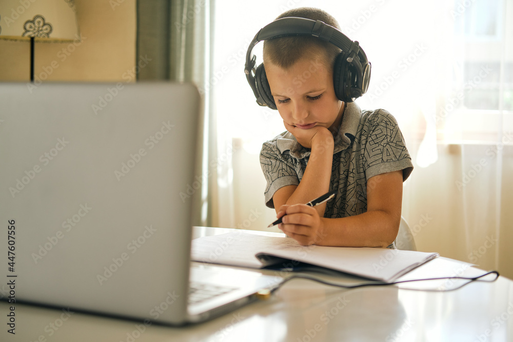 School boy in headphones writing in notebook and using laptop while doing schoolwork at home or during online lesson. Online education concept. School boy in headphones writing in notebook and using laptop while doing schoolwork at home or during online lesson. Online education concept.