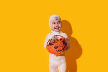 Child in mummy costume holding basket of chocolates in front of yellow background