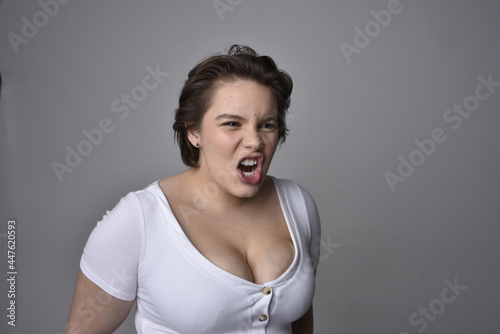 Close up portrait of young plus sized woman with short brunette hair, wearing a white shirt, with over the top emotional facial expressions against a light studio background. 