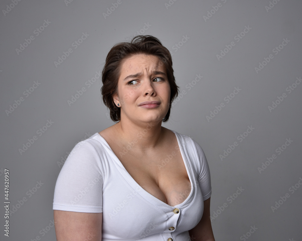 Close up portrait of young plus sized woman with short brunette hair,  wearing a white shirt, with over the top emotional facial expressions against a light studio background.  