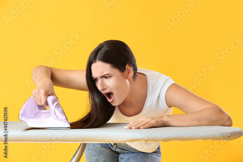 Shocked young woman ironing her hair on color background