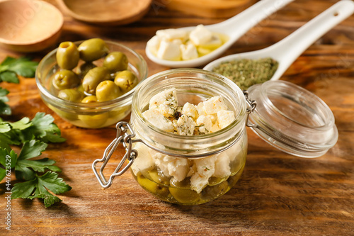 Glass jar with tasty feta cheese, oil, olives and spices on wooden background, closeup