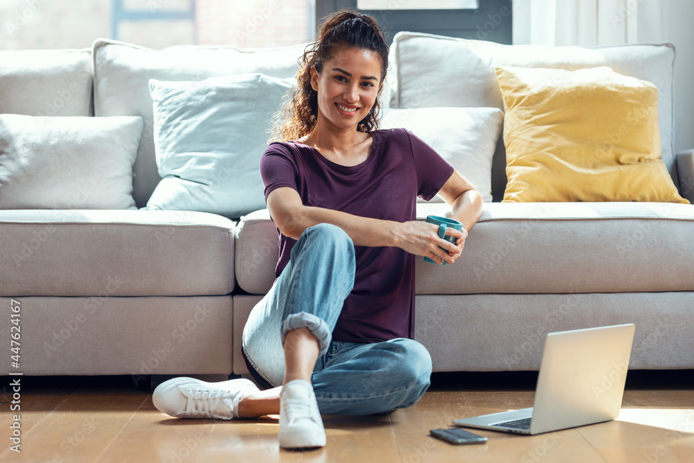 Pretty young woman using her laptop while drinking a cup of coffee sitting on the floor at home. Pretty young woman using her laptop while drinking a cup of coffee sitting on the floor at home.