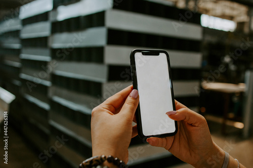A hand holds a white mobile phone with an empty white screen on the background of the factory