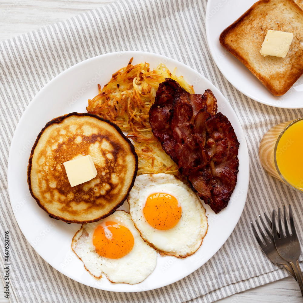 Full American Breakfast with Bacon, Hash Browns, Eggs and Pancakes on a plate on a white wooden table, top view. Flat lay, overhead, from above. Full American Breakfast with Bacon, Hash Browns, Eggs and Pancakes on a plate on a white wooden table, top view. Flat lay, overhead, from above.