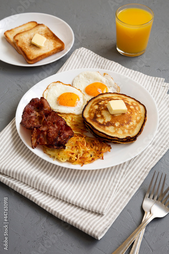 Full American Breakfast with Bacon, Hash Browns, Eggs and Pancakes on a plate on a gray surface, low angle view.