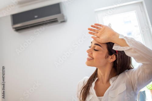 Young woman adjusts the temperature of the air conditioner using the remote control in room at home. Portrait Of A Happy Woman Holding Remote Control In Front Of Air Conditioner At Home