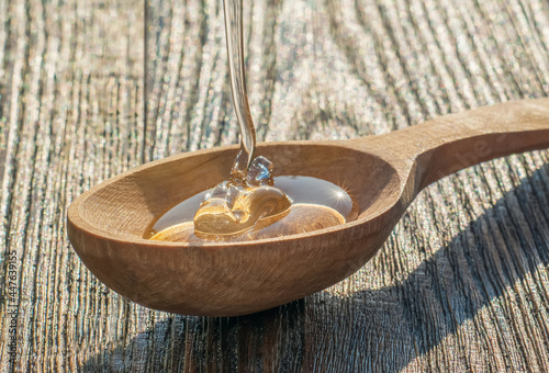 A wooden spoon in which honey is poured, while he sparkles from the rays of the sun. A spoon lies on a textured old table.