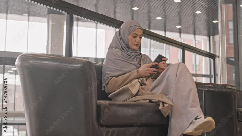 PAN slowmo shot of contemporary Arabic woman in hijab scrolling on smartphone while sitting on leather armchair at comfortable lounge zone of modern office building PAN slowmo shot of contemporary Arabic woman in hijab scrolling on smartphone while sitting on leather armchair at comfortable lounge zone of modern office building