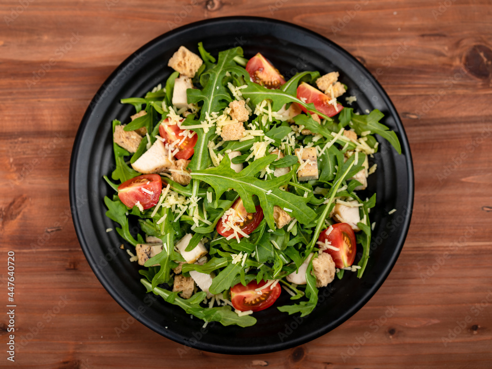 Fresh rocket salad with tomato, crispy crouton, small pieces of herb roasted chicken and parmesan cheese in black ceramic dish on dark wooded table Fresh rocket salad with tomato, crispy crouton, small pieces of herb roasted chicken and parmesan cheese in black ceramic dish on dark wooded table