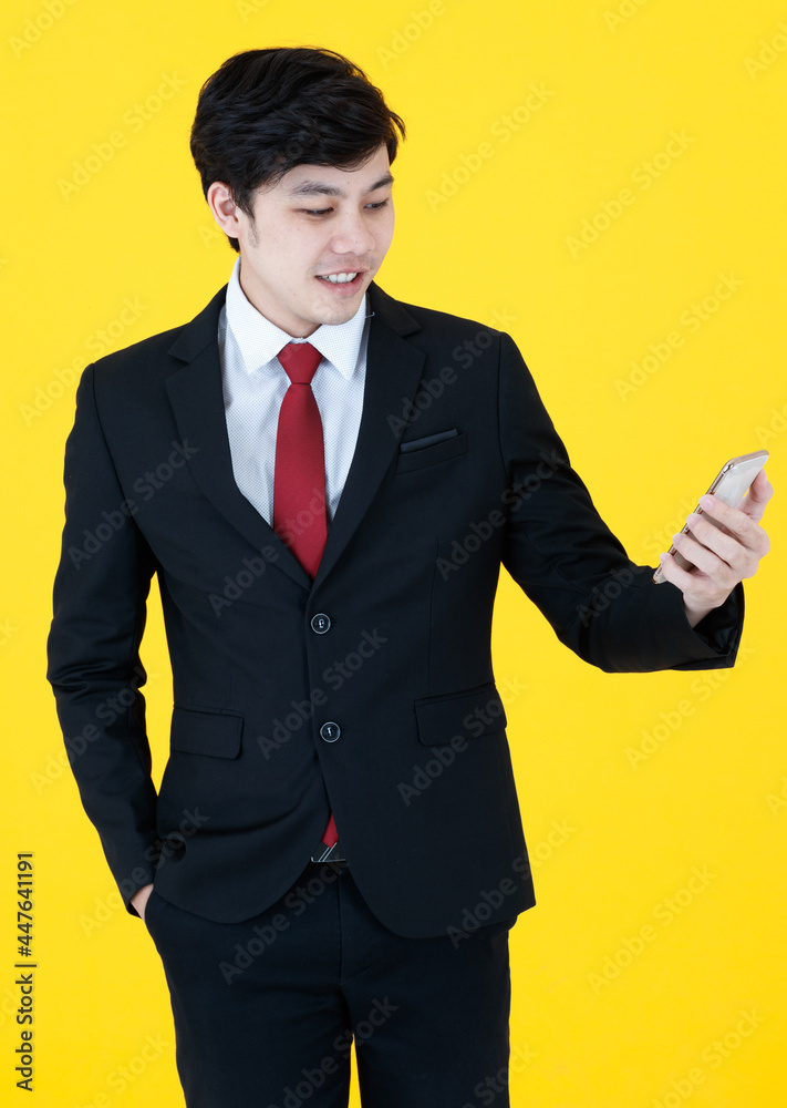 Vertical portrait shot of young adult Asian handsome man in a black long sleeve suit holding a smartphone with hand in pocket posing isolated with yellow background in studio. Concept of technology Vertical portrait shot of young adult Asian handsome man in a black long sleeve suit holding a smartphone with hand in pocket posing isolated with yellow background in studio. Concept of technology