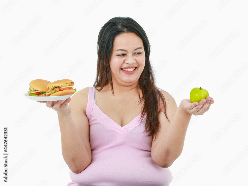 Isolated studio shot of Asian happy big fat overweight girl smiling determining choosing healthy organic green apple to eat over temptation unhealthy hamburgers in front of white background Isolated studio shot of Asian happy big fat overweight girl smiling determining choosing healthy organic green apple to eat over temptation unhealthy hamburgers in front of white background