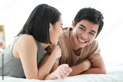 Cute smiling young Asian lover couple lying on the bed, looking at each other while talking funny in the bedroom at home. Concept of happy boyfriend and girlfriend living together