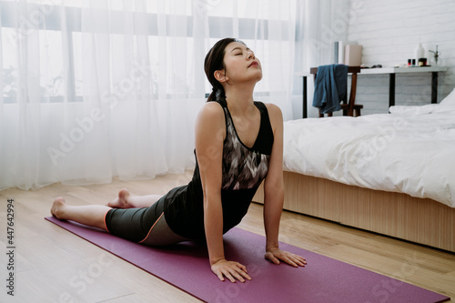 indoor recreation asian woman doing yoga to releases stress in bedroom. chinese girl practicing cobra pose by straightening arms to lift the chest off the floor, closing eye and taking in deep breath
