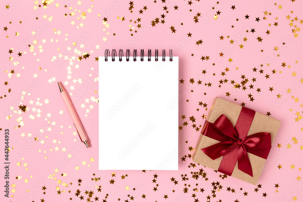 Blank notebook mockup, gift box with red ribbon and gold stars confetti on a pink background. Blank notebook mockup, gift box with red ribbon and gold stars confetti on a pink background.