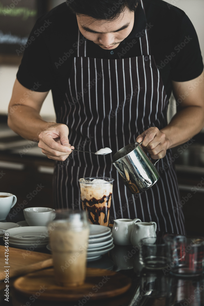 Young Asian barista man on black-and-white apron using spoon to deliberately topping coffee with milk froth for softness and mellowness at counter surrounding by dishes, jars, glass Young Asian barista man on black-and-white apron using spoon to deliberately topping coffee with milk froth for softness and mellowness at counter surrounding by dishes, jars, glass