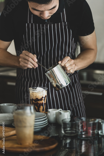Young Asian barista man on black-and-white apron using spoon to deliberately topping coffee with milk froth for softness and mellowness at counter surrounding by dishes, jars, glass