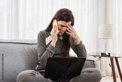 Young beautiful Brazilian woman using smartphone and working with laptop while sitting at sofa, working from home concept.