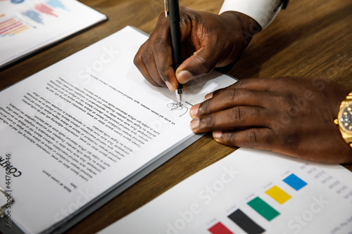 Close up and selective focus on African black formal professional business male hands holding pen, signing in white paper form or application to confirm and deal contract agreement on table.