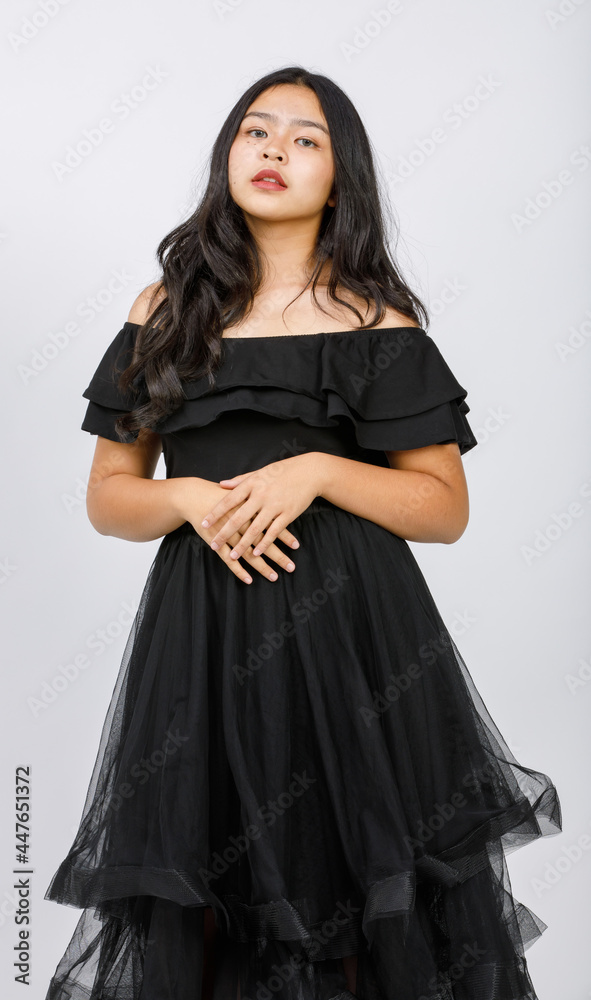 Vertical portrait shot of a cute smiling young girl teenager in a black dress looking at camera in studio isolated white background. Junior girl in fashionable clothes touching hair with shy emotion Vertical portrait shot of a cute smiling young girl teenager in a black dress looking at camera in studio isolated white background. Junior girl in fashionable clothes touching hair with shy emotion
