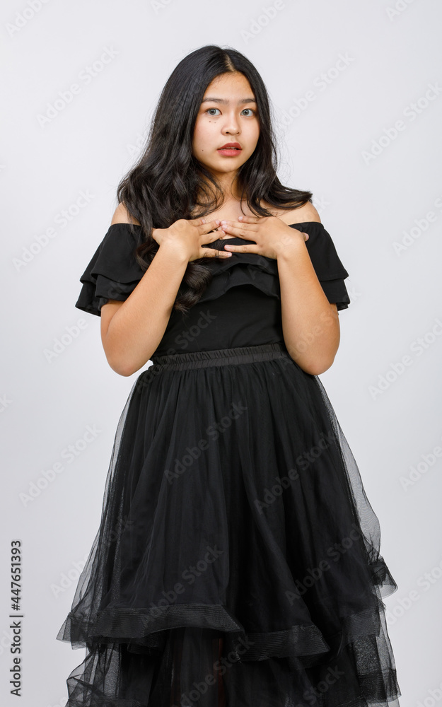Vertical portrait shot of a beautiful young girl teenager in black dress standing and looking to the camera with raising hand up to chest to showing her exciting emotion isolated on white background Vertical portrait shot of a beautiful young girl teenager in black dress standing and looking to the camera with raising hand up to chest to showing her exciting emotion isolated on white background