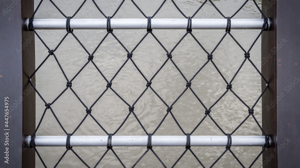 Fence decorated with black nylon net and silver rails. It is on a bridge over river.