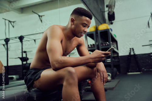 Fototapeta Naklejka Na Ścianę i Meble -  Young African American male looking at his phone to reply to an online message. Mixed race, male personal trainer looking at his phone while sitting down in the gym. High quality photo 