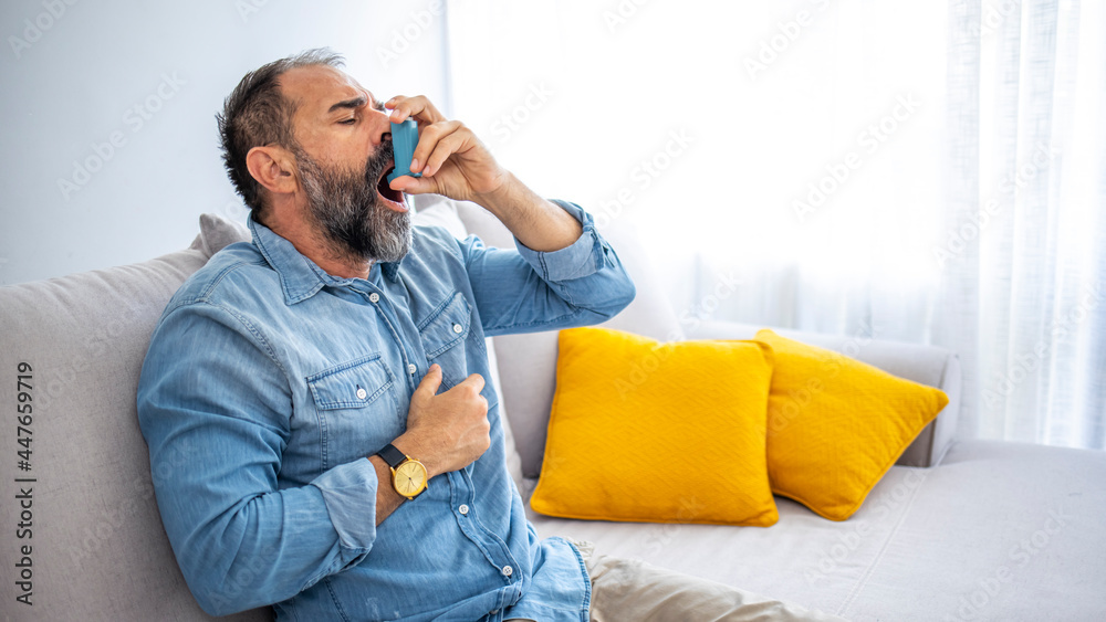 Cropped shot of a handsome mature man sitting alone at home and using an asthma pump. Man Inhaling Asthmatic Cure at Home. Mature man using medical inhaler to prevent shortness of breath