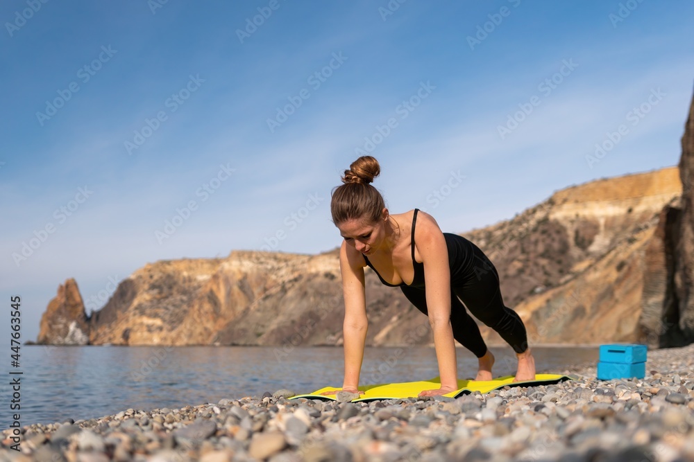 Young woman with long hair, fitness instructor in black Sportswear Leggings and Tops, stretching before pilates, on a yoga mat near the sea on a sunny day, female fitness yoga routine concept