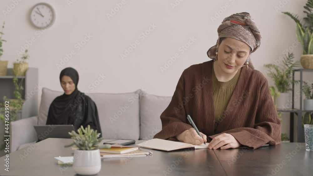 Medium slowmo shot of young attractive Muslim woman in turban taking notes in copybook at table while her female friend or colleague working on laptop sitting on sofa in background Medium slowmo shot of young attractive Muslim woman in turban taking notes in copybook at table while her female friend or colleague working on laptop sitting on sofa in background