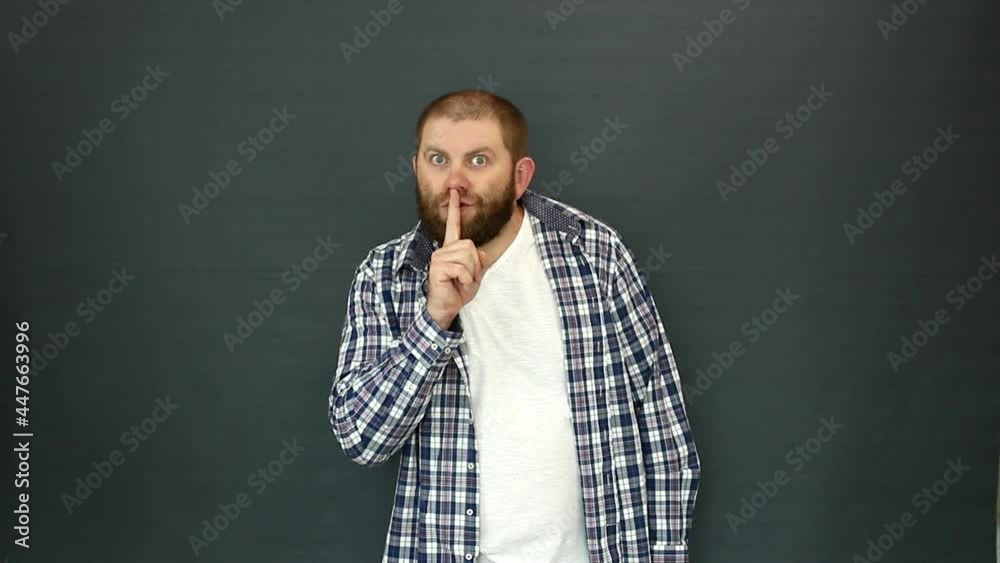 A man with a beard puts his index finger to his lips, on a gray background. The gesture indicates that you need to behave quietly. A man with a beard puts his index finger to his lips, on a gray background. The gesture indicates that you need to behave quietly.