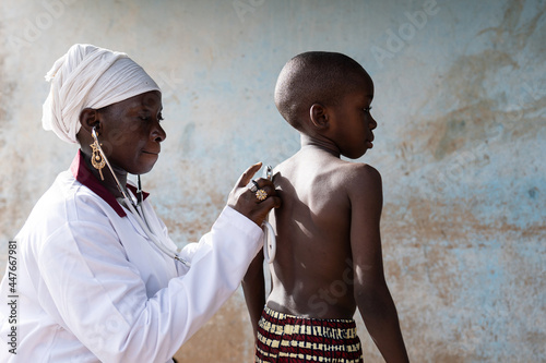 Attentive black doctor placing a stethoscope on the bare back of a standing thin schoolboy during a routine screening campaign in rural Africa