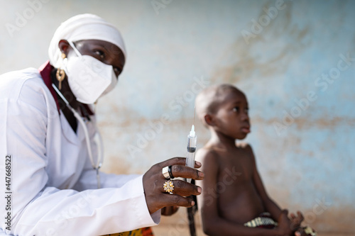 Close up of a young African nurse with an scarf wrapped around her head and a protective face mask squirting a liquid from a syringe and a small black boy photo