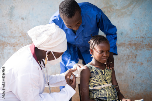 Worried little African schoolgirl during flu vaccination getting her intramuscular shot from a young black female pediatrician while beeing comforted by the doctor's colleague