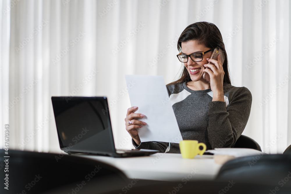 Young beautiful Brazilian woman using smartphone and working with laptop while sitting at office desk, working from home concept.