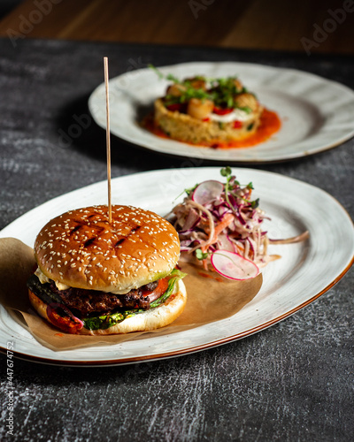 Fototapeta Naklejka Na Ścianę i Meble -  Fresh tasty burger with soft bun meat cutlet and fresh vegetables served with vegetable salad, White oval plate with whiskey on background. Vertical orientation