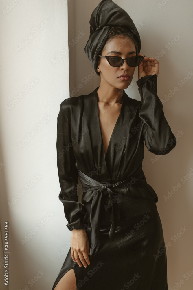 African woman in dark modern dress and sunglasses posing inside. Beautiful girl in trendy hat looking away in apartment..