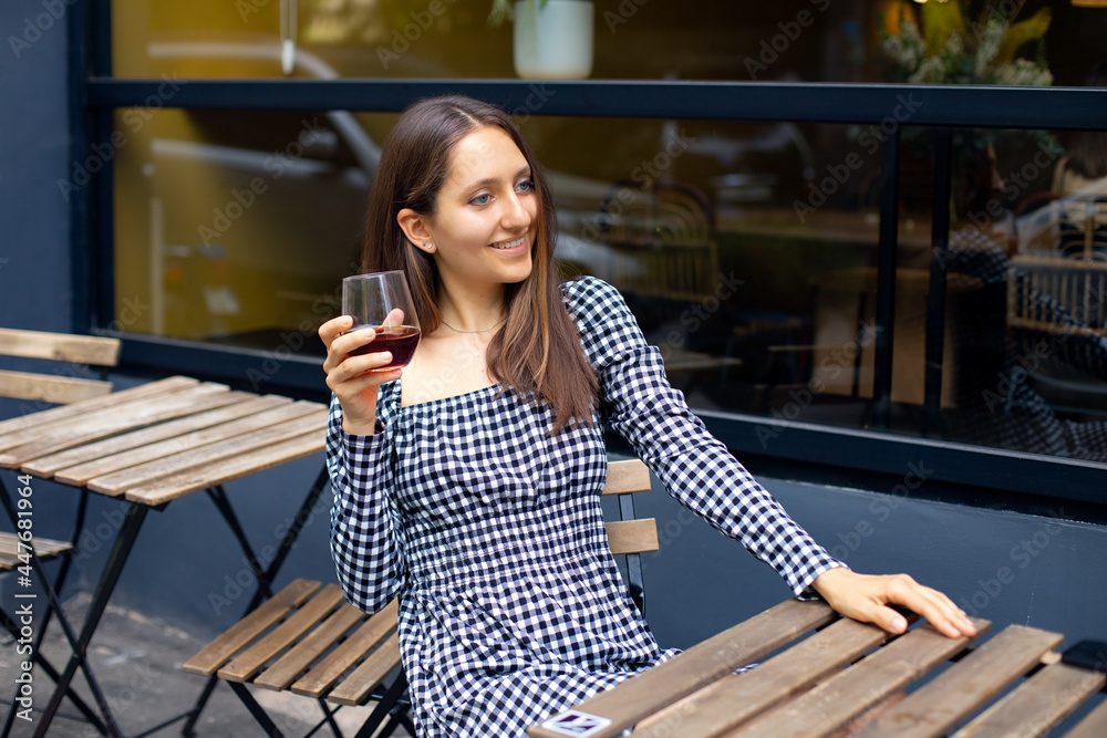 caucasian girl in plaid dress with drink smiling and chatting while sitting at the table in modern cafe outdoor