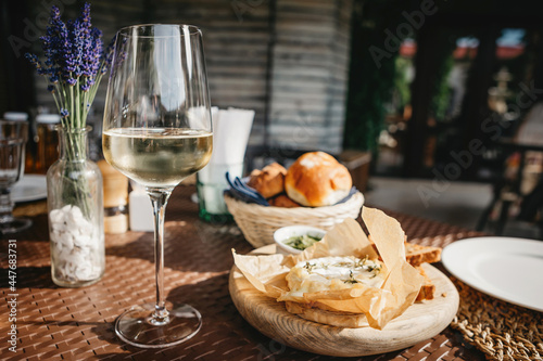 Glass of cold white wine and oven baked camembert cheese with rosemary on baking paper on wooden plate. Serving lunch at the winery, rustic style table setting