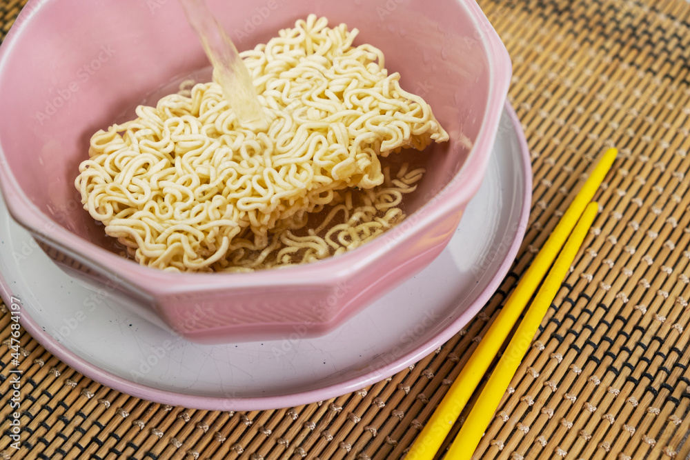 Quick instant dry noodles with pouring hot water in pink bowl on bamboo mat, Asian Chinese ramen pasta fast food eaten with yellow bamboo chopsticks