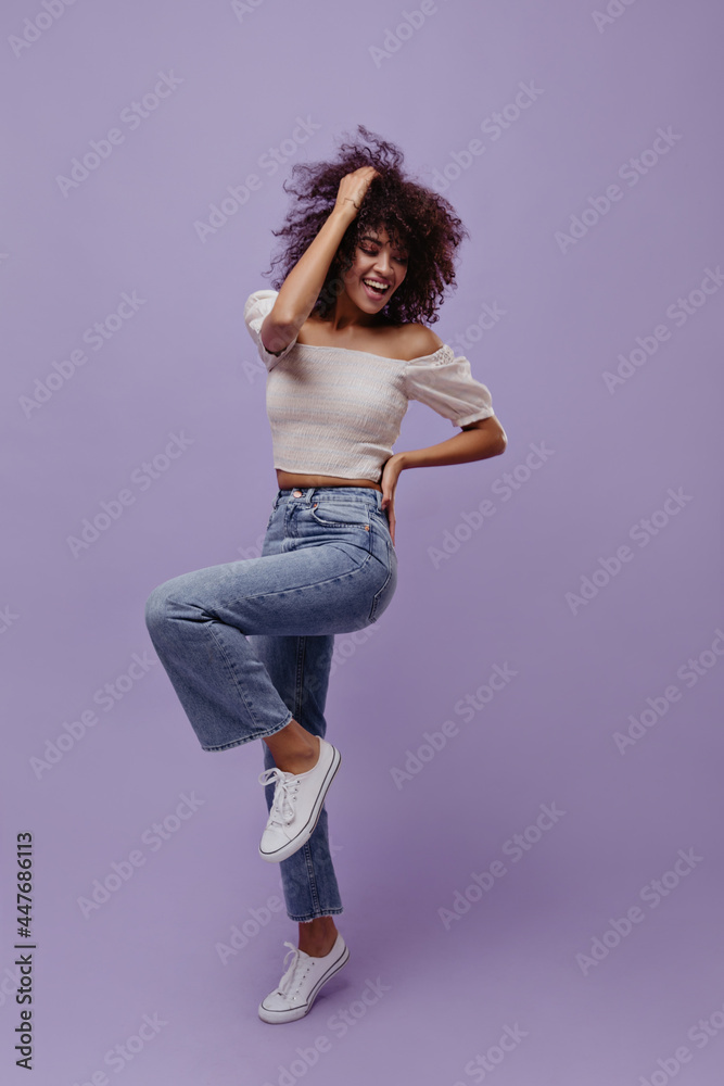 Excited brunette curly woman in jeans and cropped white top moves, smiles, touches hair and rises leg on purple background.