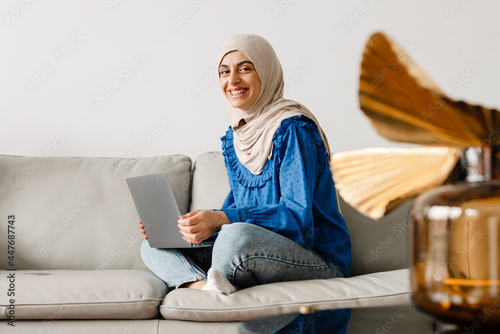 Middle eastern woman in headscarf using laptop while sitting on sofa - obrazy, fototapety, plakaty 