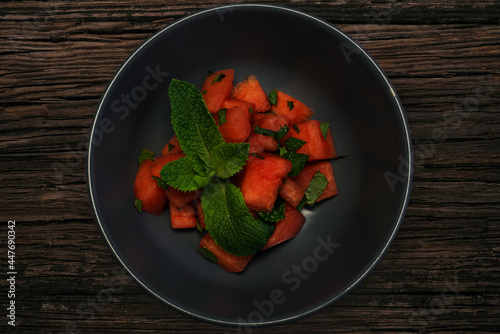 Melon pieces served with green mint leaves in a grey bowl on dark brown wooden table (ID: 447690342)
