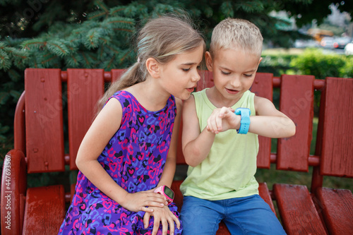 Happy kids sitting on bench and using smartwatches with interest, data synchronization between smartphone and smartwatch, new technology for children, digital education and care, outdoors lifestyle